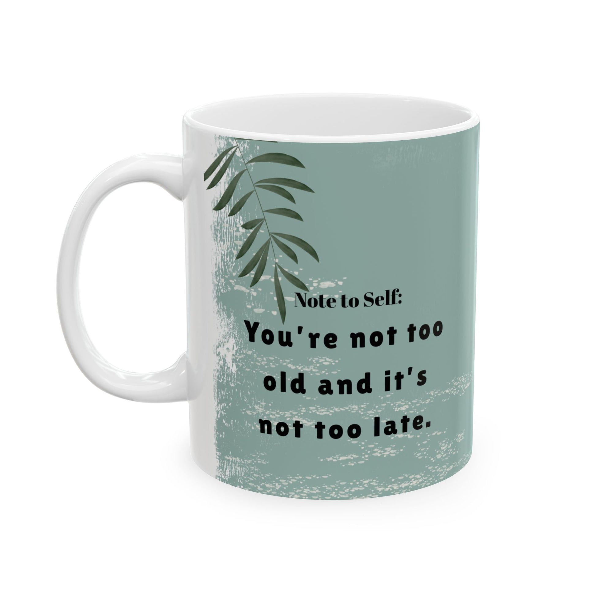 A white ceramic mug with a handle, featuring a sage green background on the right side with a motivational quote, and a white textured background with a green leafy design on the left side. The quote reads "Note to Self: You're not too old and it's not too late." The mug is positioned against a plain white background