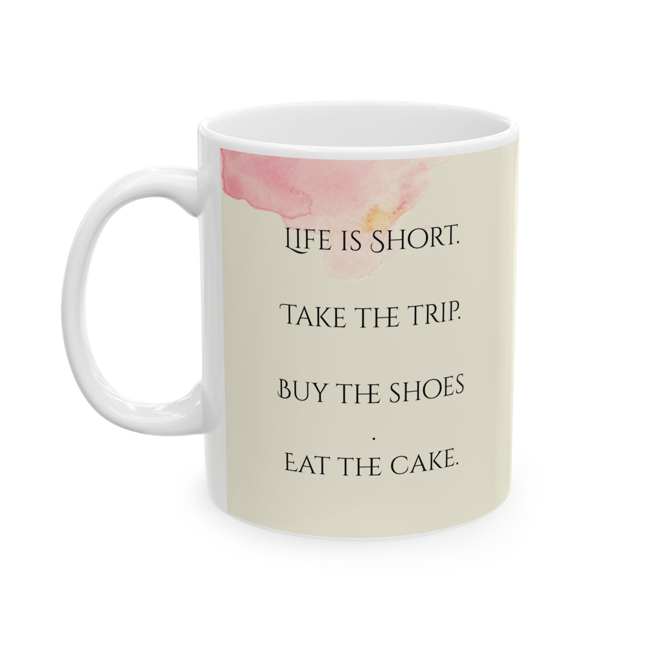 A white ceramic mug with a handle is depicted, featuring a beige background and a watercolor-style pink splotch on the left side. The mug displays the following text in black, stacked lines: "LIFE IS SHORT. TAKE THE TRIP. BUY THE SHOES. EAT THE CAKE." This text serves as an inspirational quote. The mug is positioned centrally against a plain white backdrop.