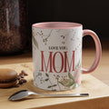 "Love You, Mom" ceramic coffee mug. The mug is white with a pink rim and handle, and it features a floral design with the text "LOVE YOU, MOM" in red lettering. A macaron, coffee beans, and a spoon are placed on the wooden surface next to the mug.