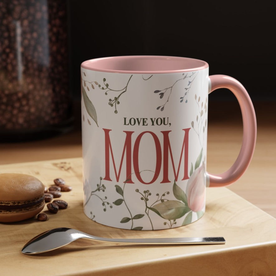"Love You, Mom" ceramic coffee mug. The mug is white with a pink rim and handle, and it features a floral design with the text "LOVE YOU, MOM" in red lettering. A macaron, coffee beans, and a spoon are placed on the wooden surface next to the mug.